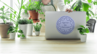 A periwinkle 6-inch circular sticker with a dark blue abstract line drawing of a circle of people with joined arms surrounding the hand written words Together We Survived. It is on the back of a silver laptop surrounded by green, indoor potted plants
