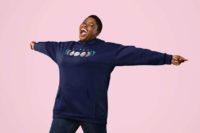 7 phases of the moon in a gradient of the transgender pride colors: blue, pink, and white. It is demoed on the front of a navy pullover hoodie on a plus size black woman, her arms stretched wide, looking ecstatic.
