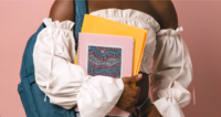 A 6 by 8 inch square sticker with a design of a detailed mosaic of abstract waves in the colors of the transgender pride flag, pink, baby blue, and white. A black gender nonconforming person is holding a pink notebook with the sticker on the front.