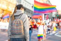 A brunette woman in a hoodie and baseball cap at a pride parade wearing a drawstring bag with a repeat pattern of abstract waves in the colors of the transgender pride flag, with a detailed mosaic texture. It has two black cords for shoulder straps.