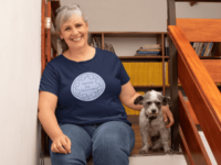 An abstract line drawing of a circle of people with joined arms surrounding the hand written words Together We Survived in dark blue on a periwinkle circle. It is on a navy T-shirt worn by a happy, plus size, middle aged woman sitting with her dog.