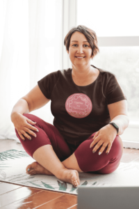 An abstract line drawing of a circle of people with joined arms surrounding the hand written words Together We Survived in white on a dark pink circle. It is on a dark brown T-shirt worn by a plus sized middle aged woman sitting on a yoga mat.